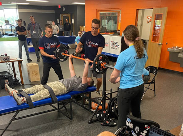 Person lying on bench lifting a weight bar with spotters on each side.